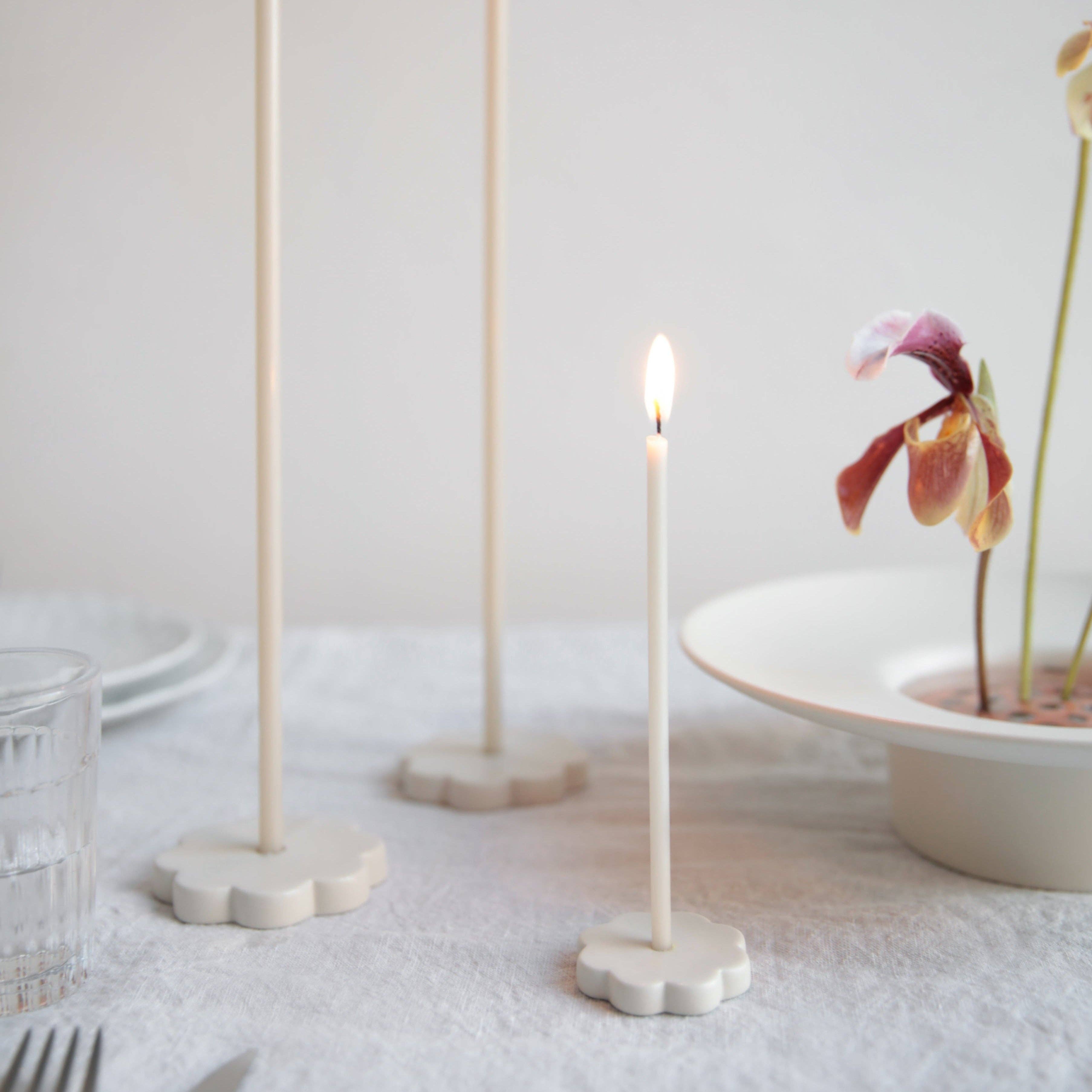 Three minimalist cream-colored ceramic candle holders with flower-shaped bases are arranged on a white tablecloth. One holder is lit with a small flame, and a white plate with a dried orchid is visible in the softly blurred background.