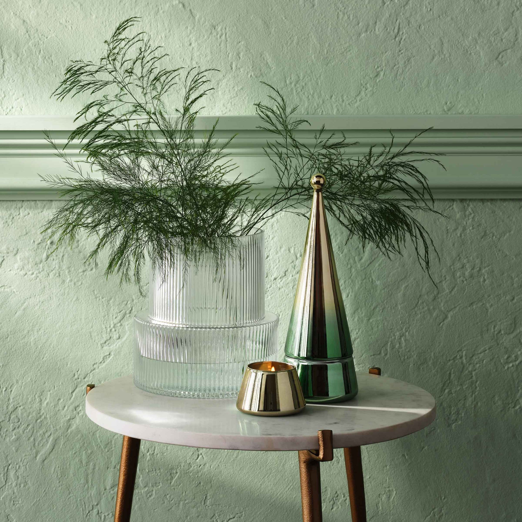 Small round table with a plant, metallic vase, and candle against a light green wall.