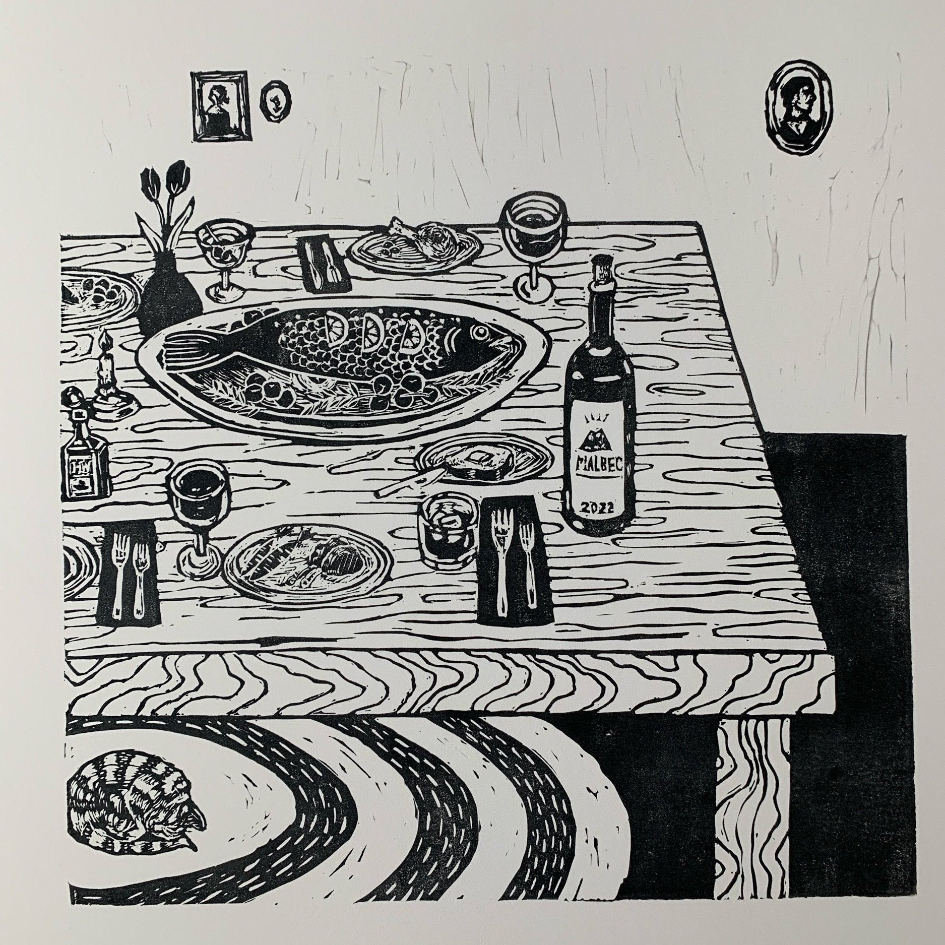 Black-and-white relief linocut of a wooden dining table set for a meal: a large whole fish platter at center, plates, forks, wine glasses, small dishes, a vase of flowers, and a bottle labeled “Malbec 2022.” Framed portraits hang on the wall behind the table, and a curled cat sleeps on a patterned rug beneath the table’s edge.