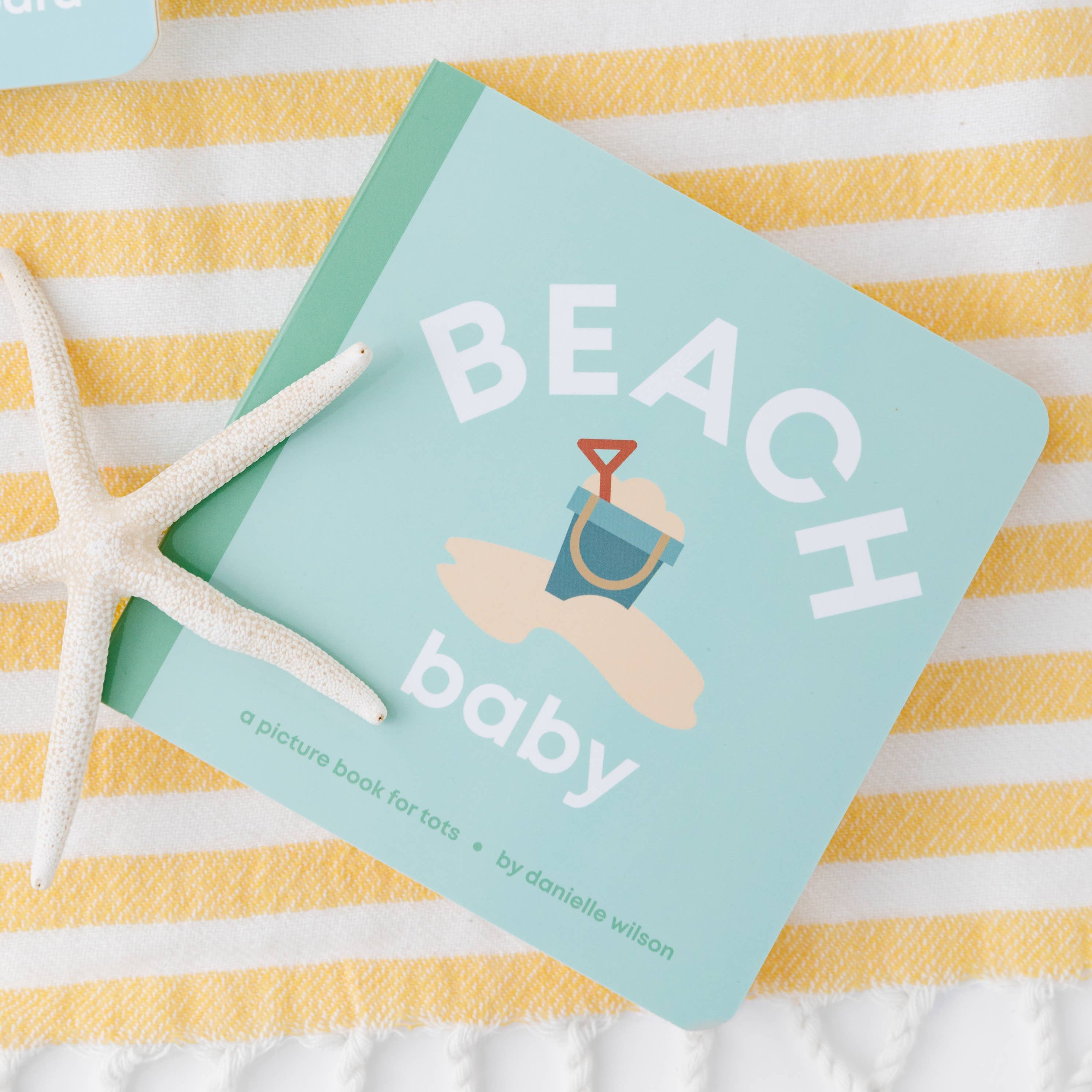Book titled 'Beach Baby' on a yellow and white striped towel with sunglasses and a starfish.