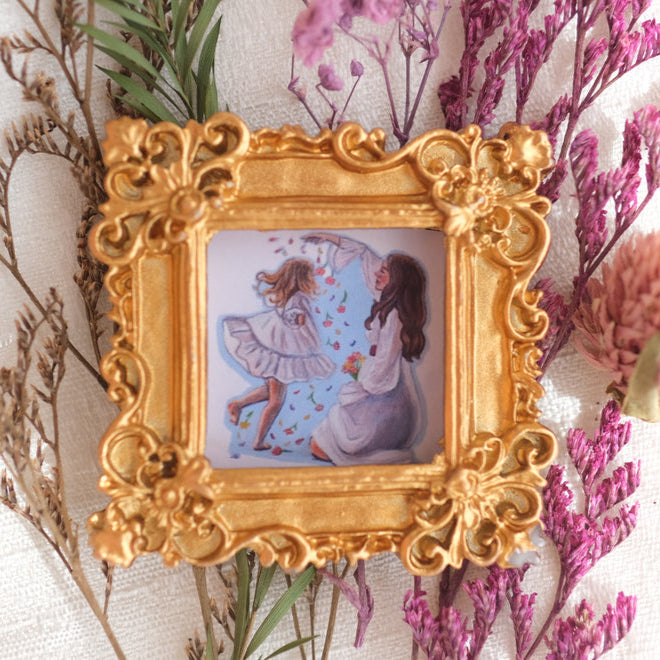 A detailed gold square frame holds a painting of a woman and a young girl joyfully playing together, with the girl in mid-dance and petals floating around them. The artwork is set against a pale blue background and is surrounded by sprigs of dried flowers on a textured fabric.