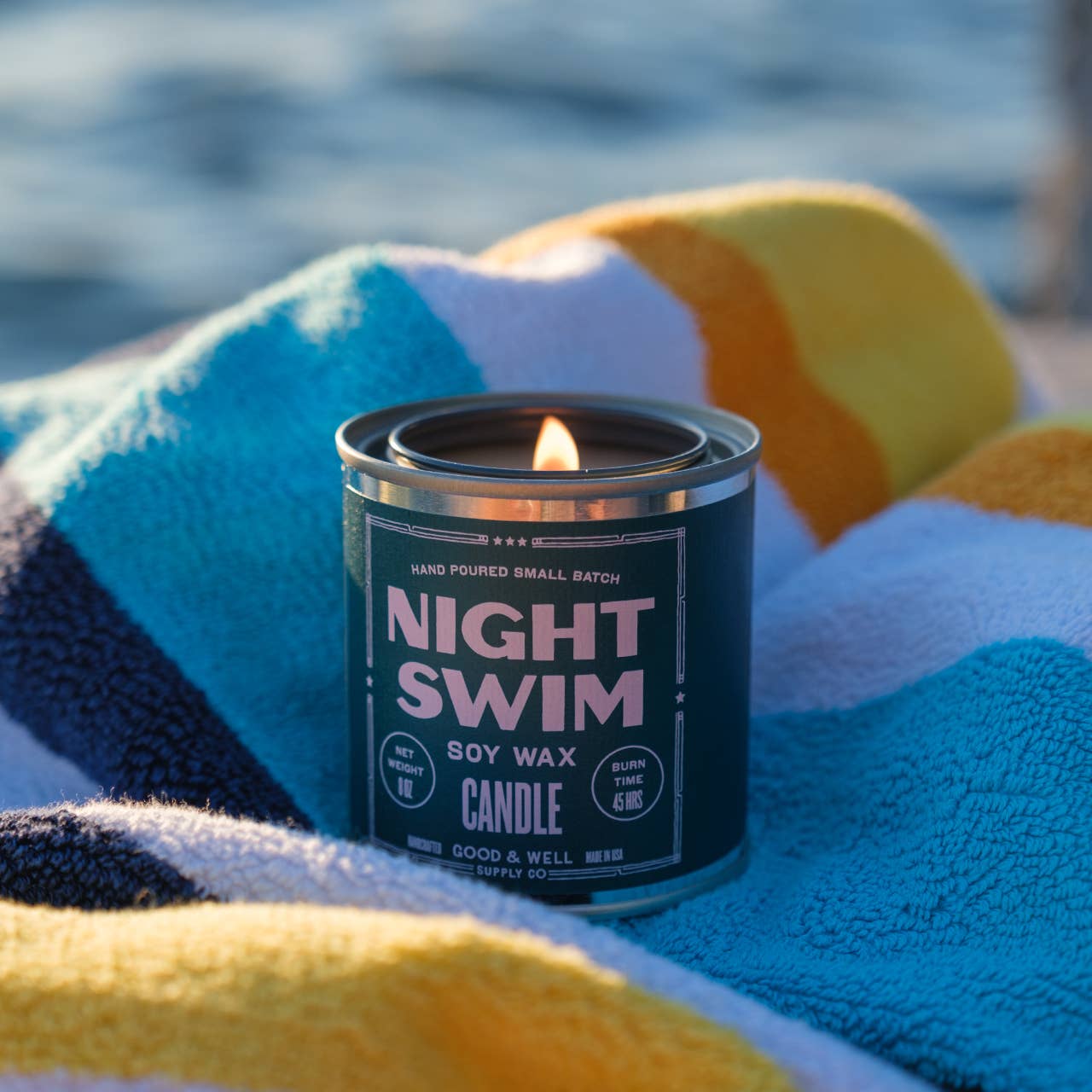Candle labeled 'Night Swim' on a striped towel by a body of water