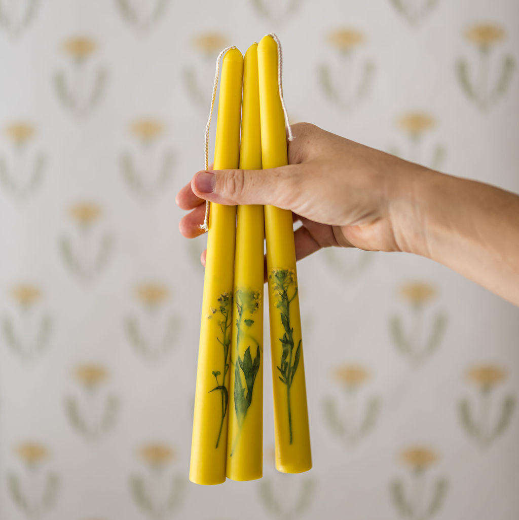 a hand holding three taper floral candles in front of a floral wallpaper background