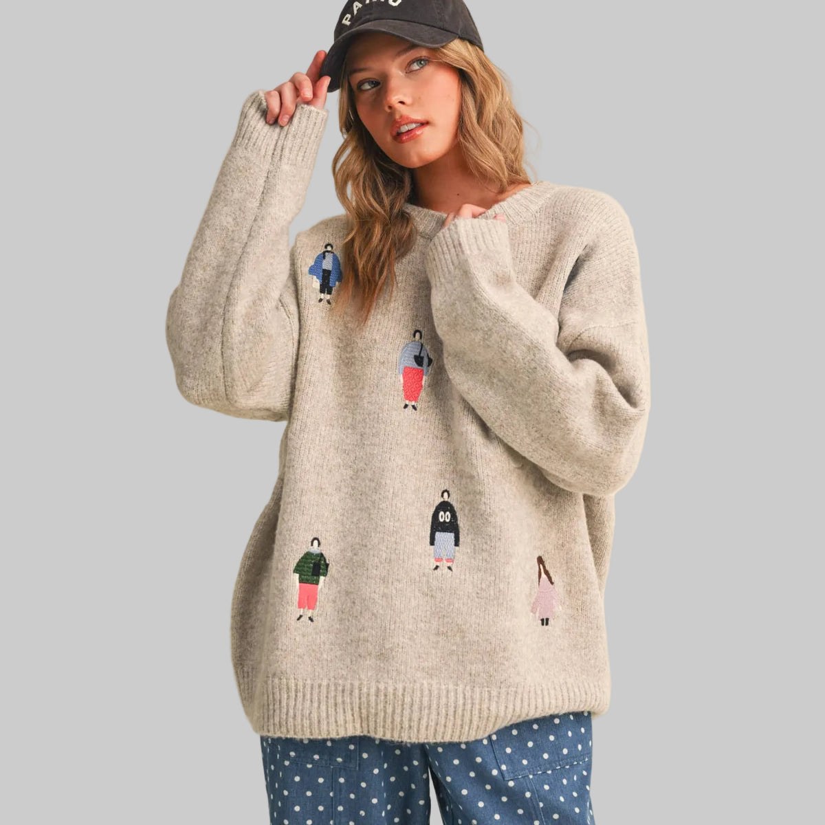 A woman wearing a beige knit sweater with small colorful women dressed in fun clothing patches, paired with gray polka-dot pants, against a plain beige studio background.
