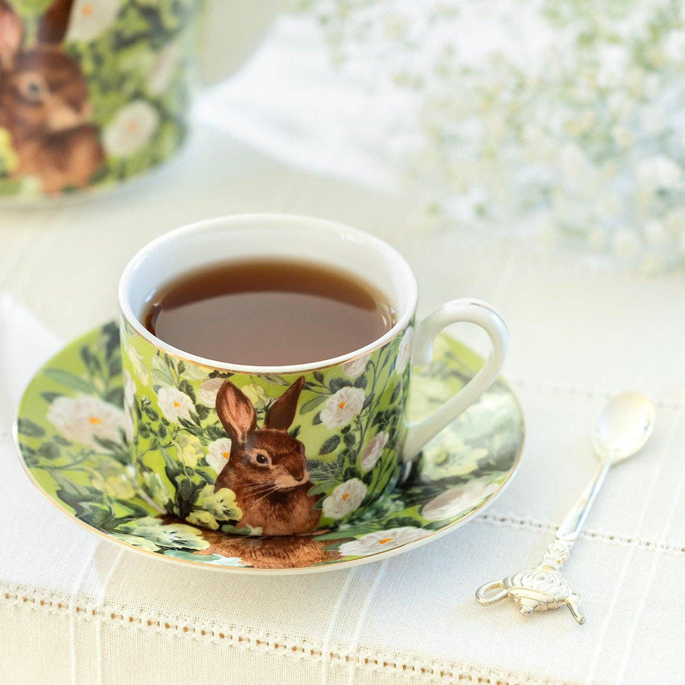 Teacup with rabbit design on a saucer, filled with tea, on a decorative tablecloth.
