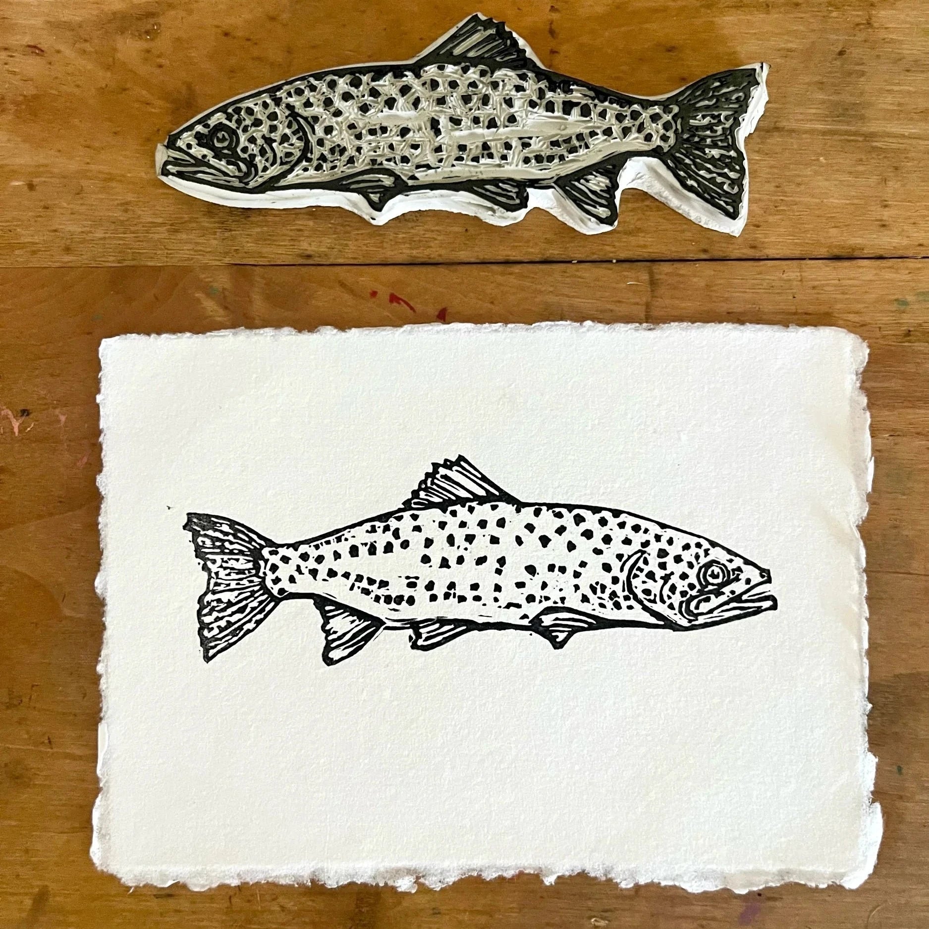 Rustic white paper with an ink impression of a spotted rainbow trout in black ink. Pictured against a wooden back drop with the fish stamp situated above the print.