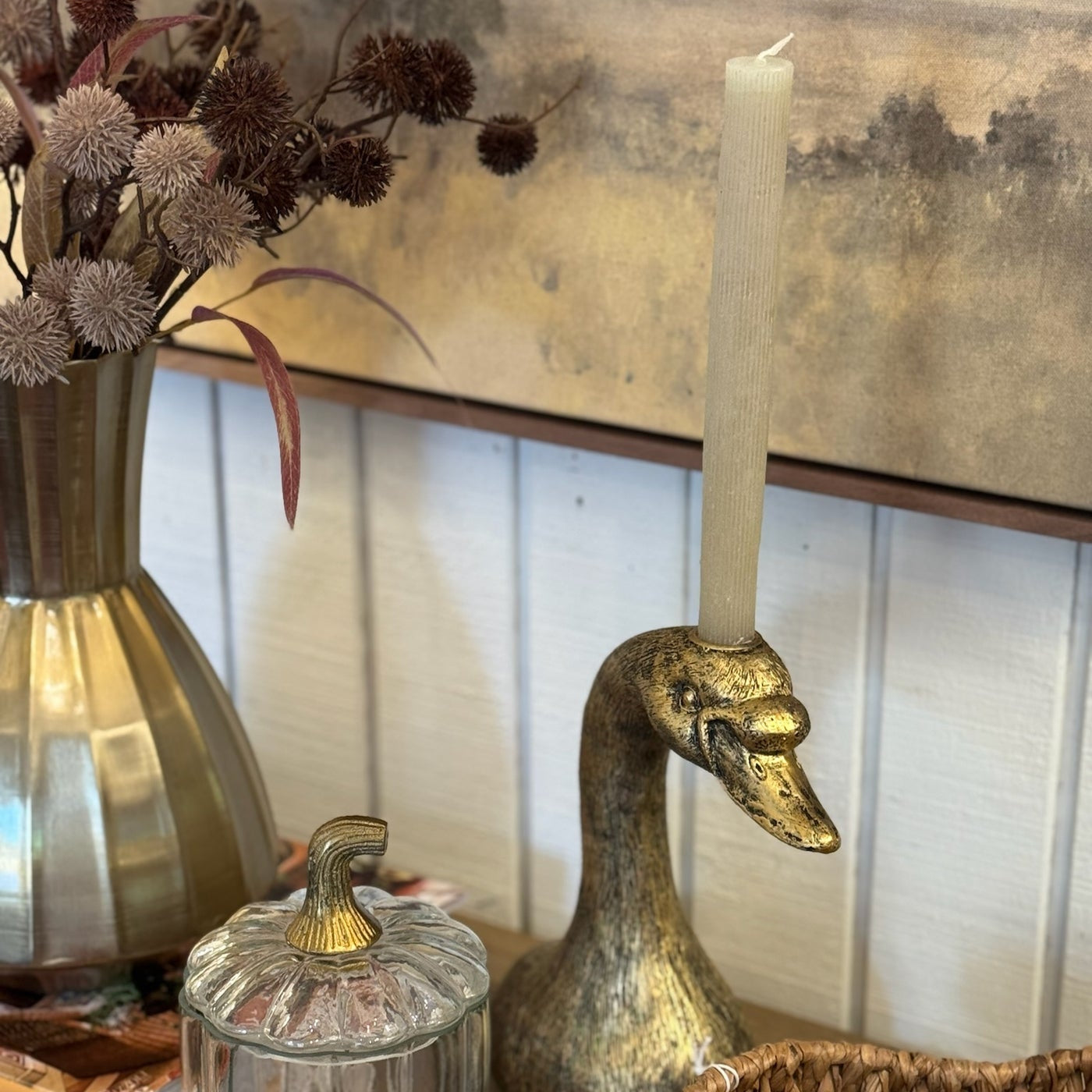 Decorative setting with a gold duck-shaped candle holder, glass jar, and vase with dried flowers on a wooden surface.