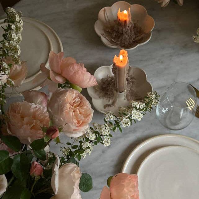 A styled tabletop scene with blush pink roses, white filler flowers, plates, a clear glass, and two lit candles in flower-shaped holders on a light marble surface.