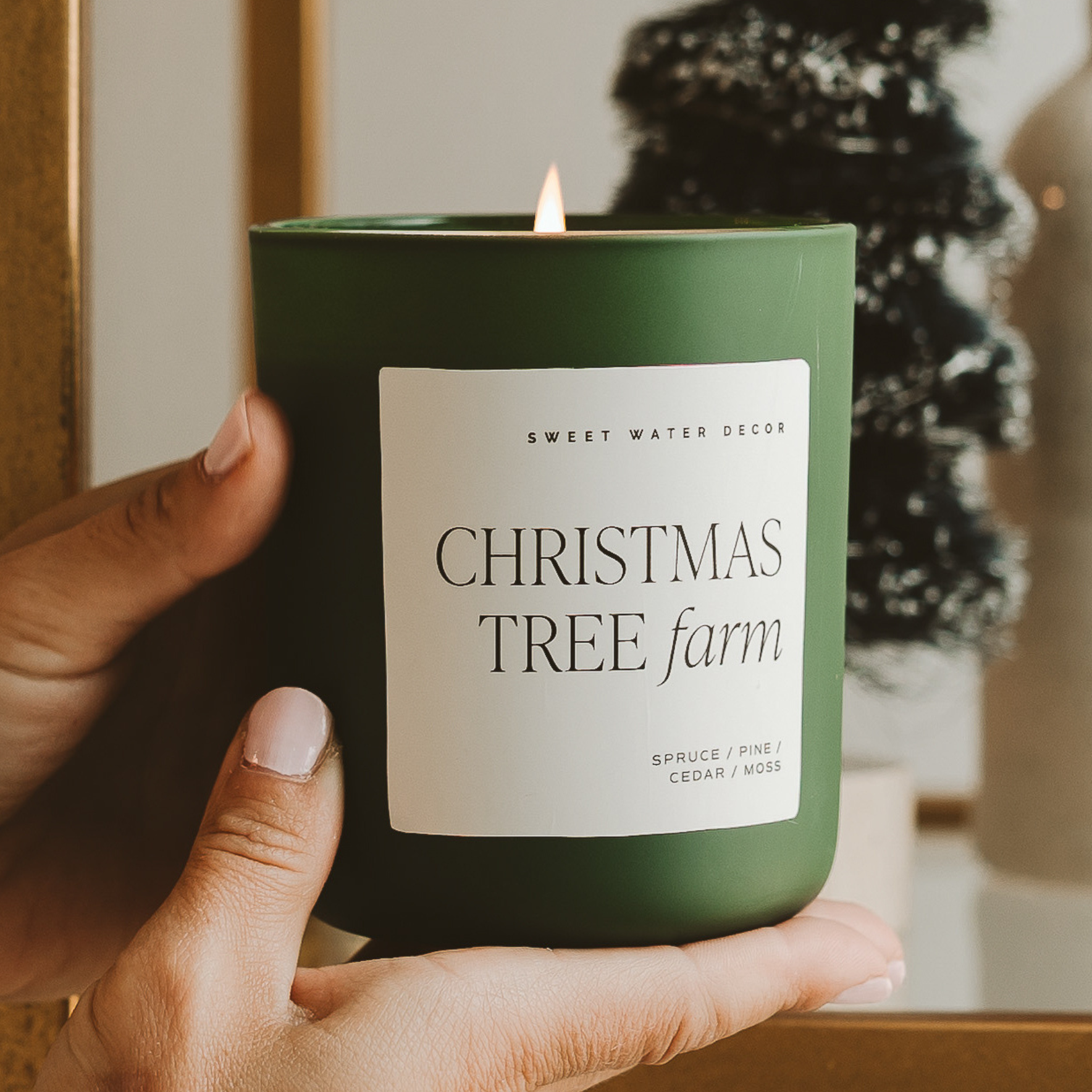 Hand holding a green candle labeled 'Christmas Tree Farm' with a blurred indoor background.