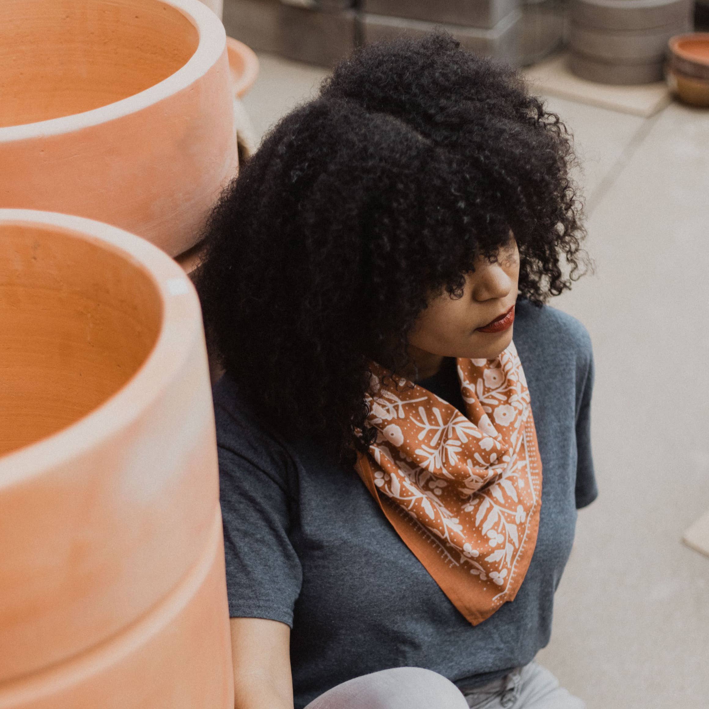 Woman with an afro wearing a patterned scarf sitting next to terracotta pots.