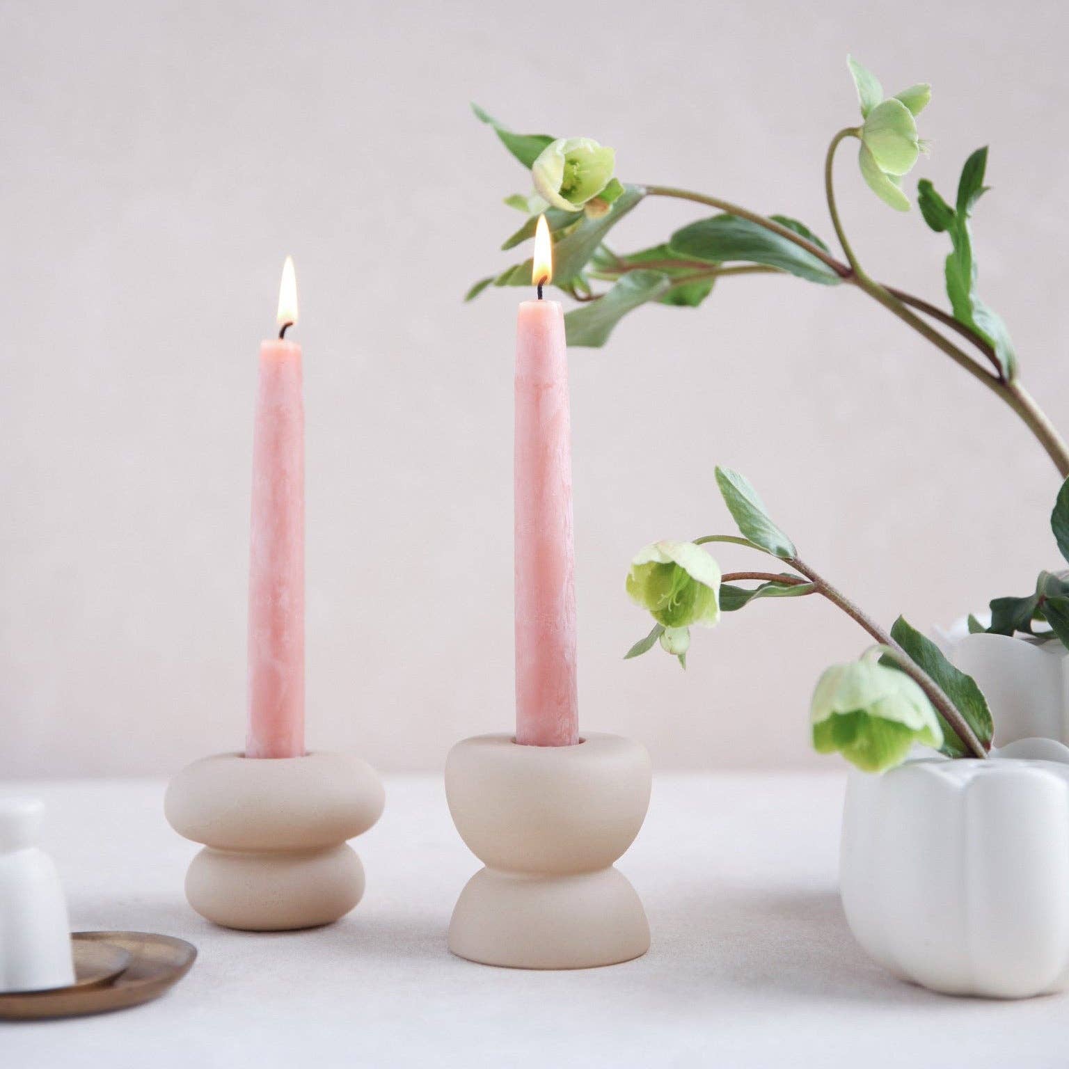 Two lit pink taper candles in matte beige stacked round holders, styled on a light surface with soft green flowers and a white vase against a pale neutral background.