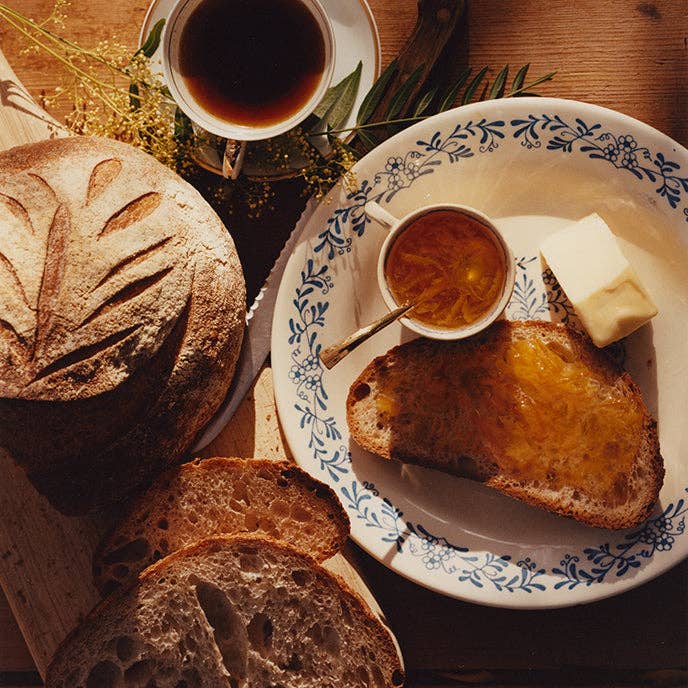 Loaf of bread with slices, a plate with toast, butter, and jam, and a cup of coffee on a wooden table.