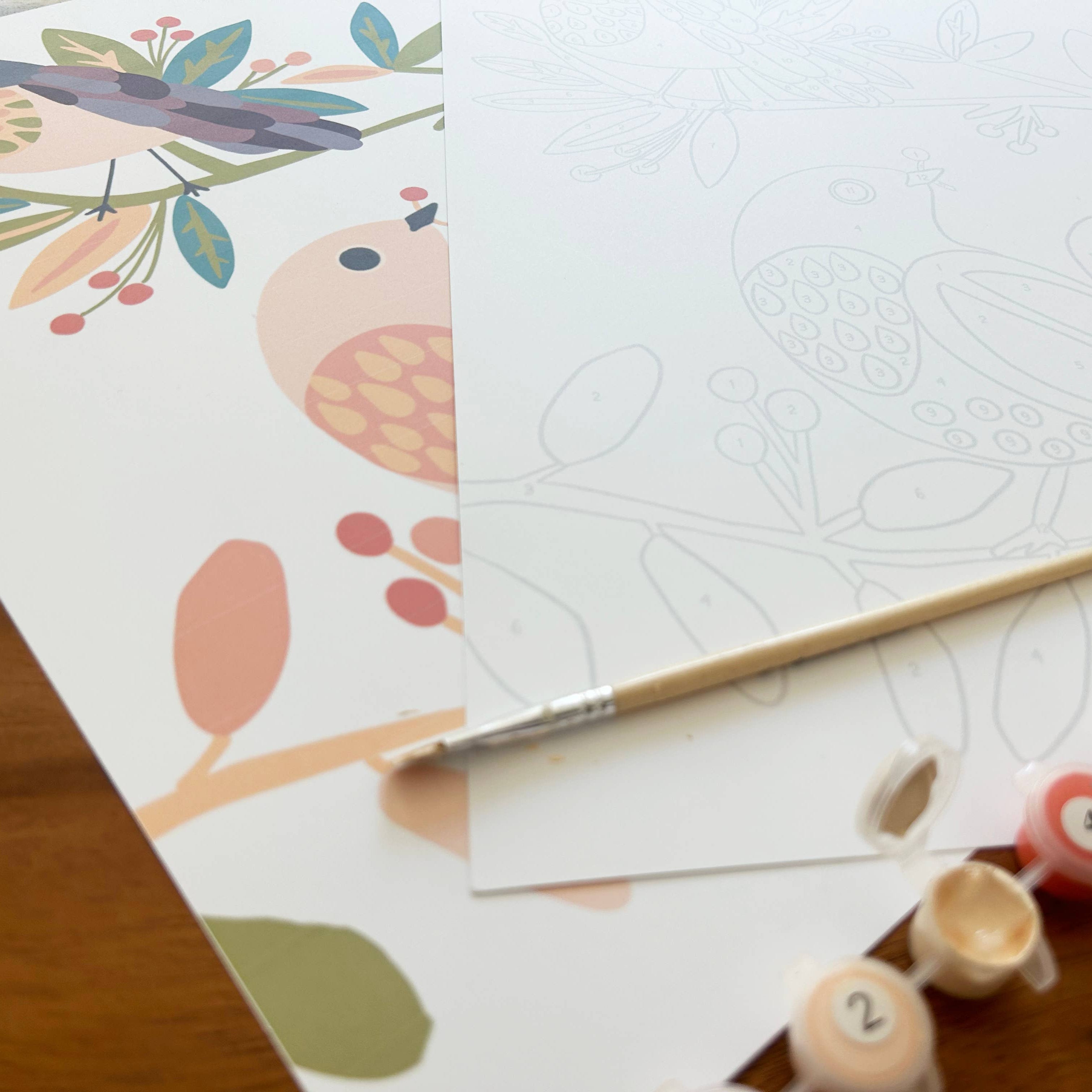 Coloring book with bird design, white paper, paintbrush, and paint tubes on a wooden surface