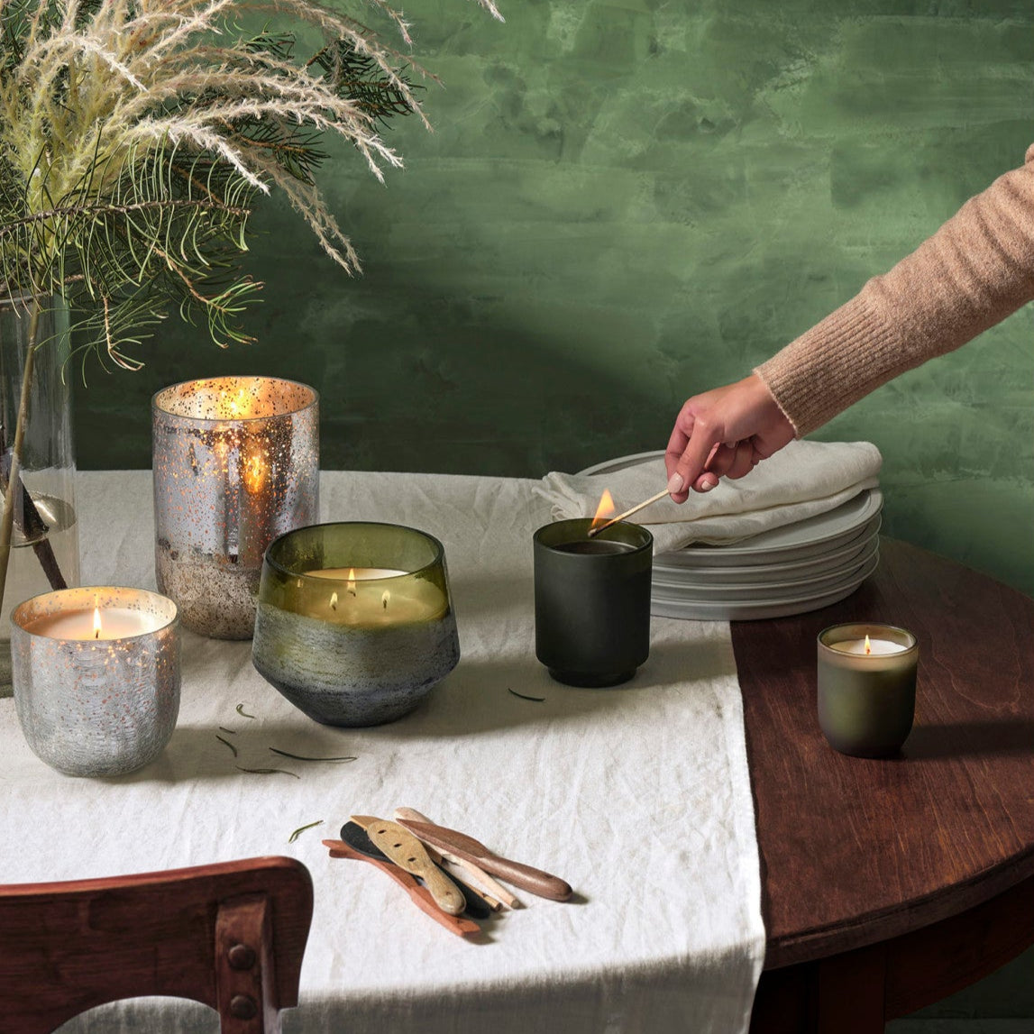 Person lighting a candle on a table with decorative elements and a green wall.