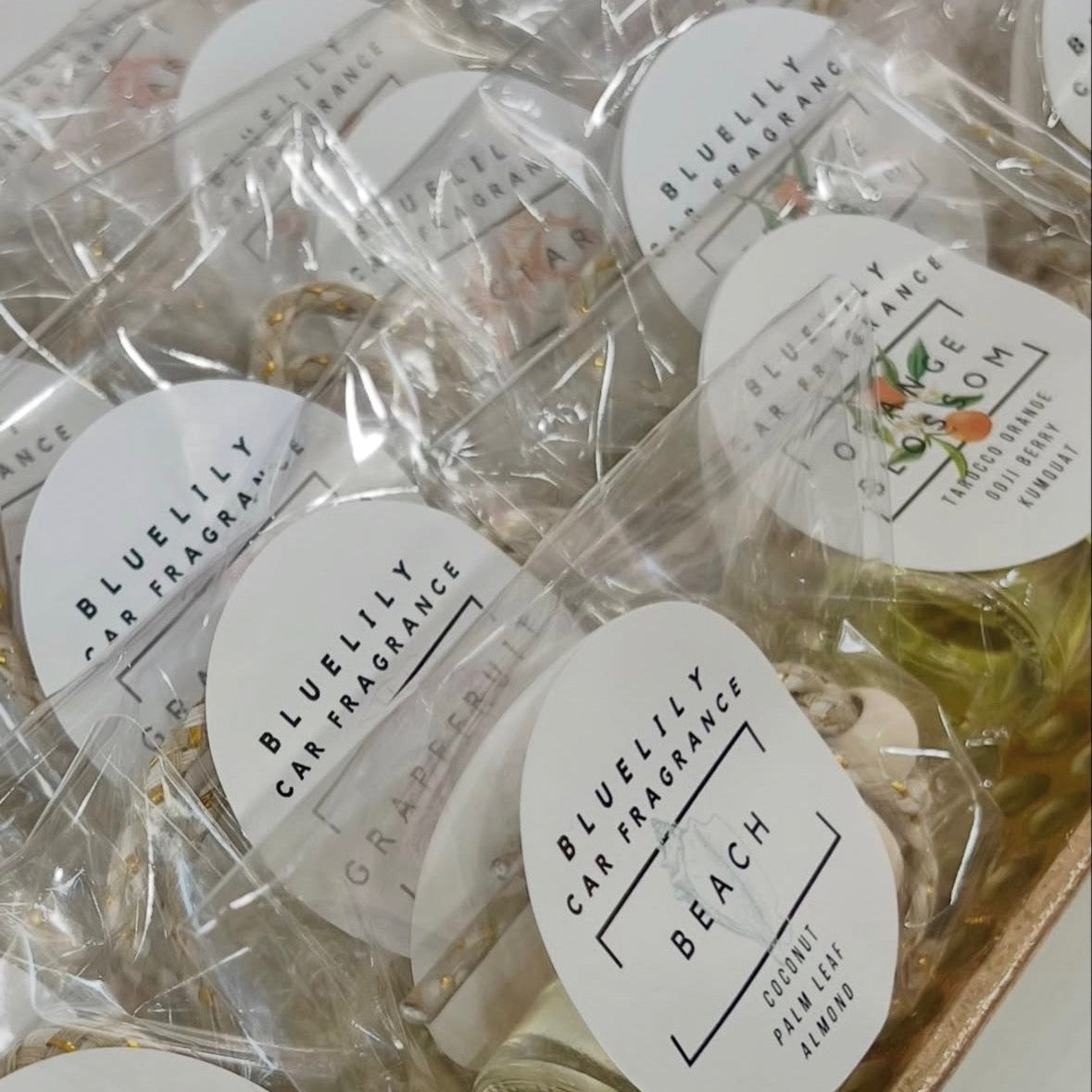 Multiple small car fragrance bottles wrapped in clear plastic with round white labels reading 'BLUELILY CAR FRAGRANCE' and scent names such as 'BEACH' and 'GRASSHOPPER', arranged in a tray.