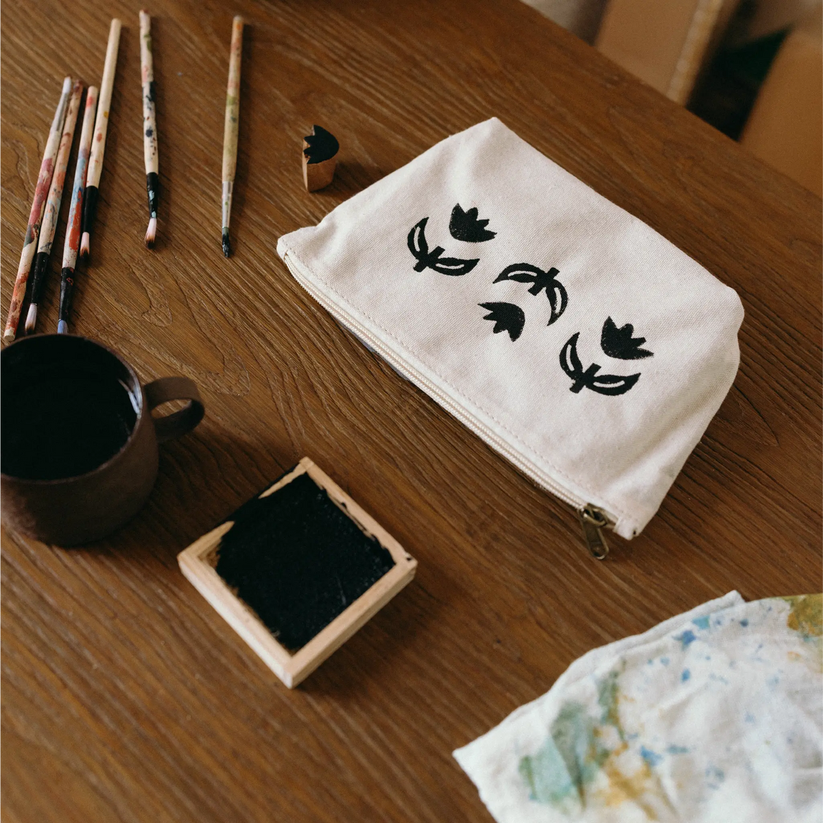A cream canvas zippered pouch with black tulip motifs, styled on a wooden table with art supplies including paintbrushes, a cup of ink, and an ink block.