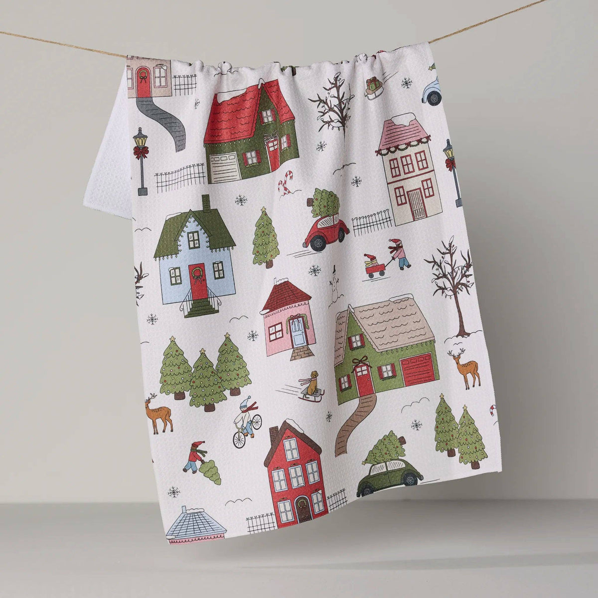 A white kitchen tea towel with a colorful winter village pattern, hanging from a string on a neutral gray background.