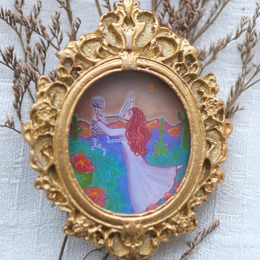 A small ornate gold frame encloses an illustration of a red-haired woman in a flowing white dress dancing joyfully with a skeleton in a colorful desert landscape filled with cacti, red flowers, and a setting sun. The frame is surrounded by delicate dried beige branches on a soft white fabric background.
