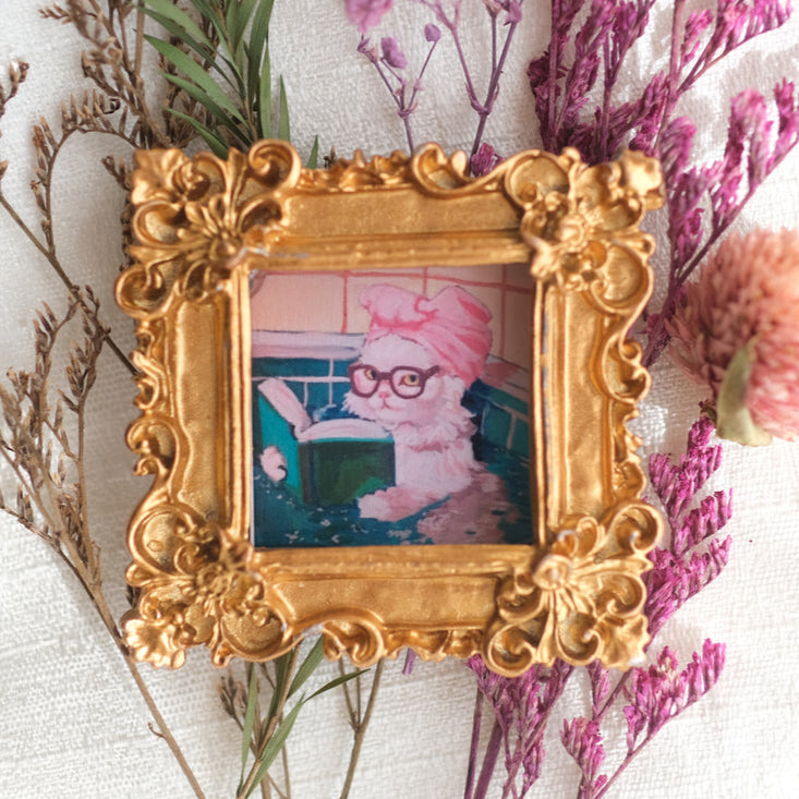 A small ornate gold frame holds a whimsical photo of a white cat wearing glasses and a pink towel on its head, sitting in a bath while reading a book. The frame is surrounded by dried flowers and greenery, giving the display a charming and vintage-inspired look.
