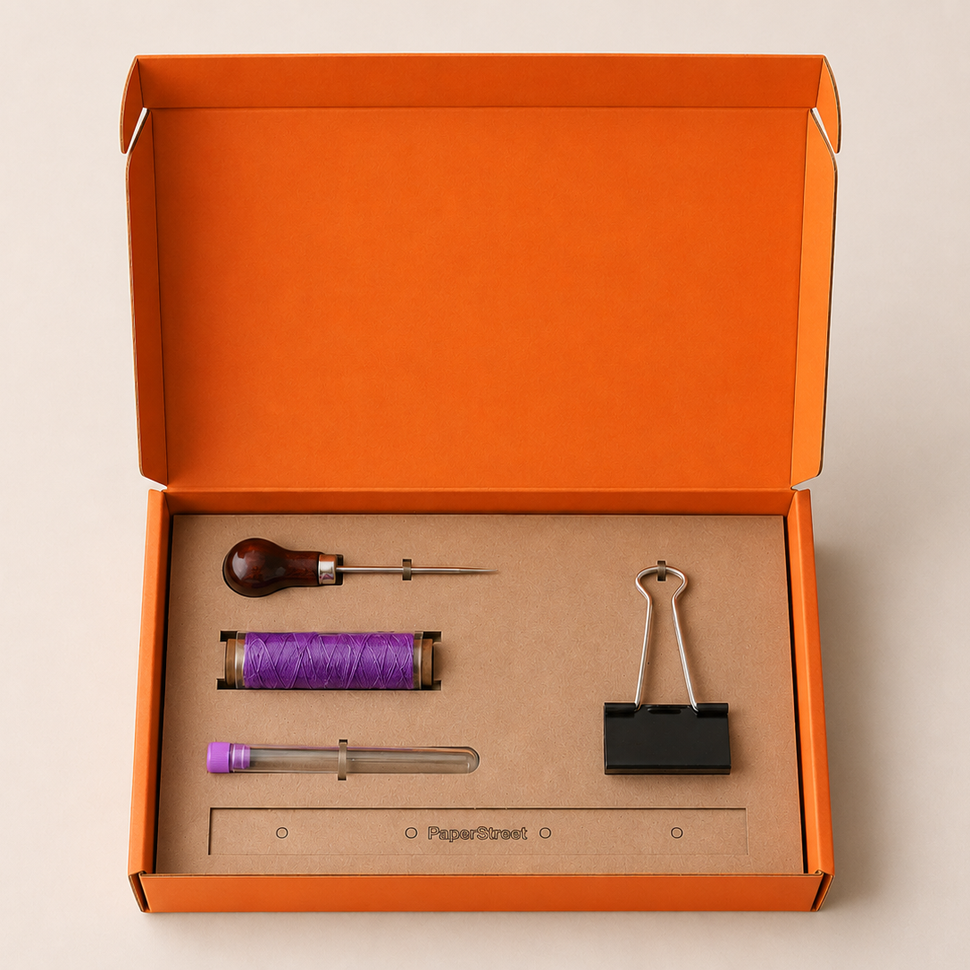 An open orange cardboard box on a light beige background containing a small sewing or craft kit with a wooden-handled needle, purple thread, a metal tool, a black binder clip, and a ruler on a brown insert.