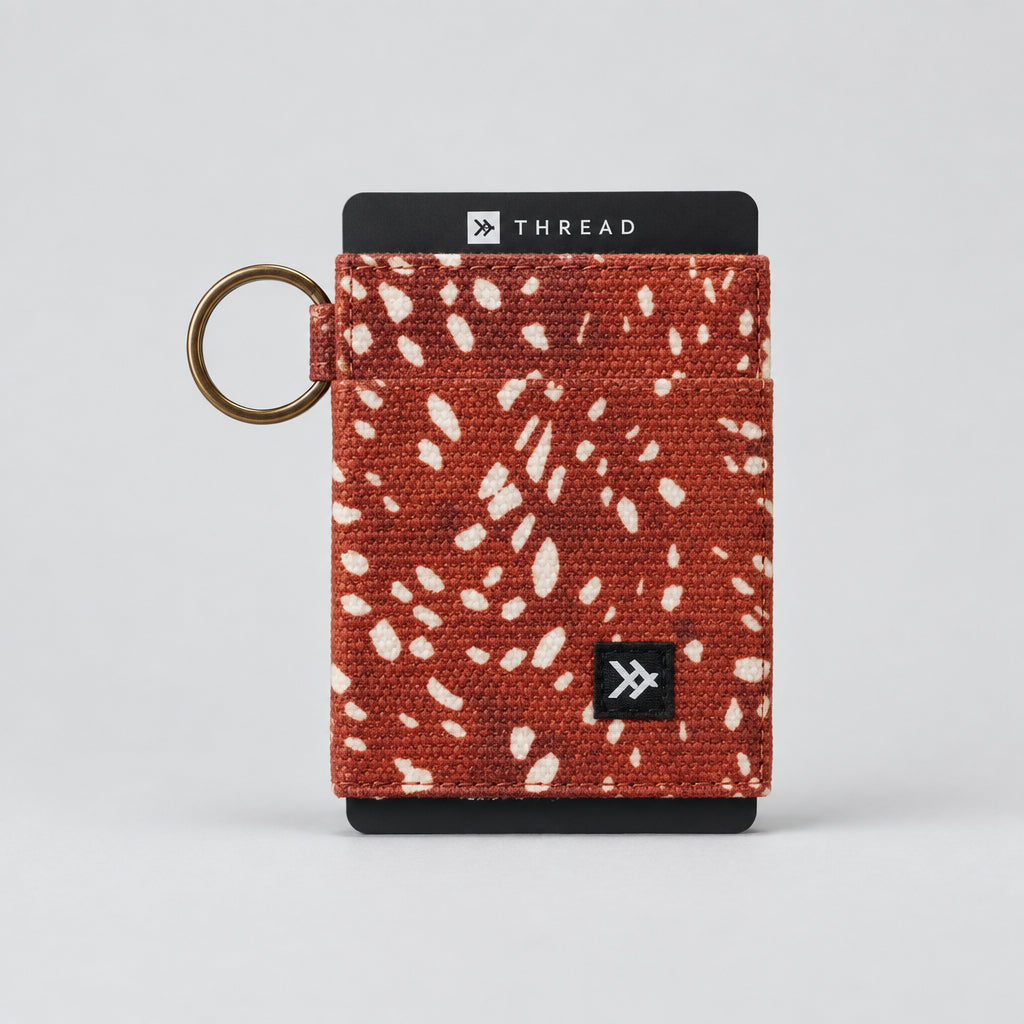 A Thread brand card holder wallet in a rust-orange woven fabric with white speckled flecks, featuring a gold-tone keyring and black end caps with branding.