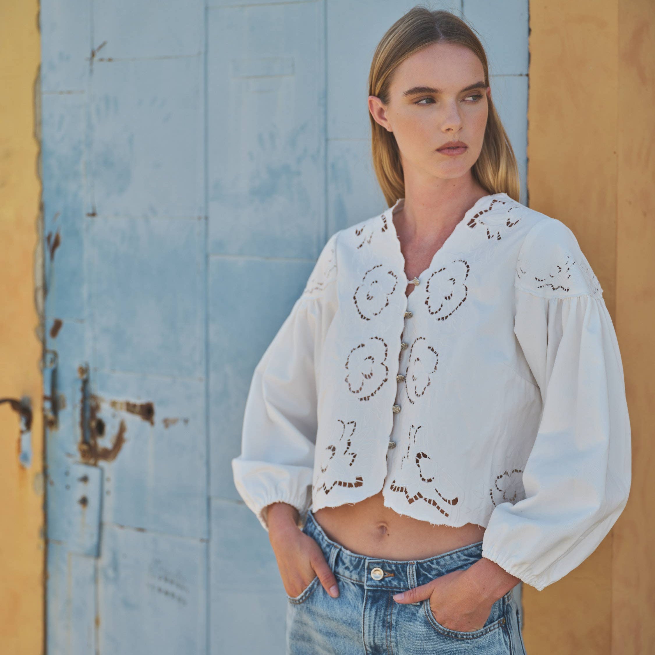 Woman wearing a white embroidered blouse and blue jeans against a rustic wall.