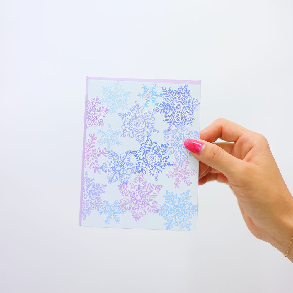 White card with whimsical purple and blue snowflakes dancing across the front. Pictured in a woman's hand against a white back ground.