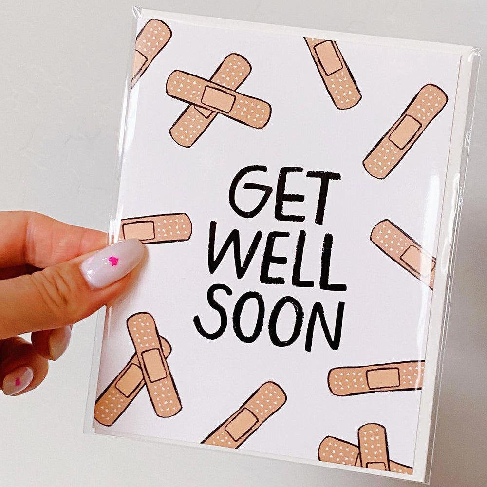 Cute bandaid design card with "Get Well Soon" printed across. Pictured in a woman's hand against a white back ground.