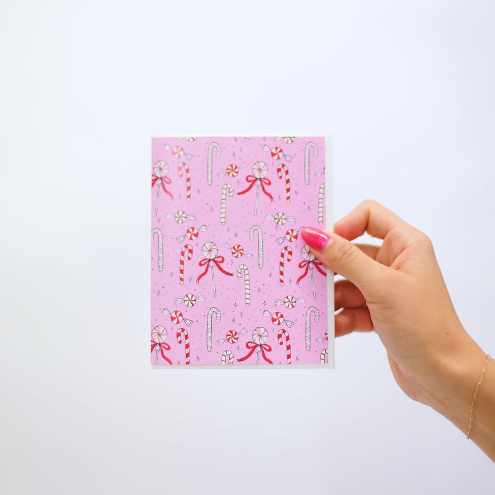 Whimsical pink card with assorted peppermint candies and bows scattered across the front. Pictured in a woman's hand against a white back ground.