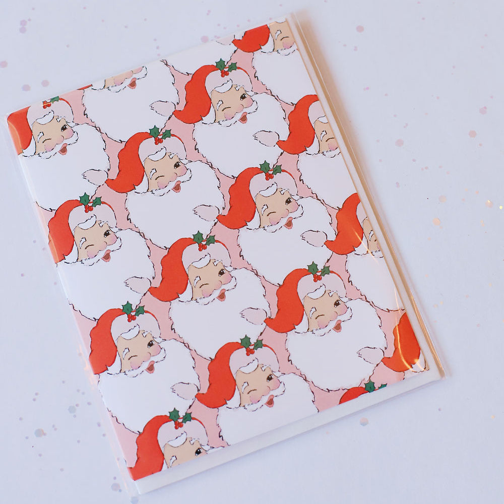 Light pink card with a classic whimsical Santa face print covering the front. Pictured against a white back ground.