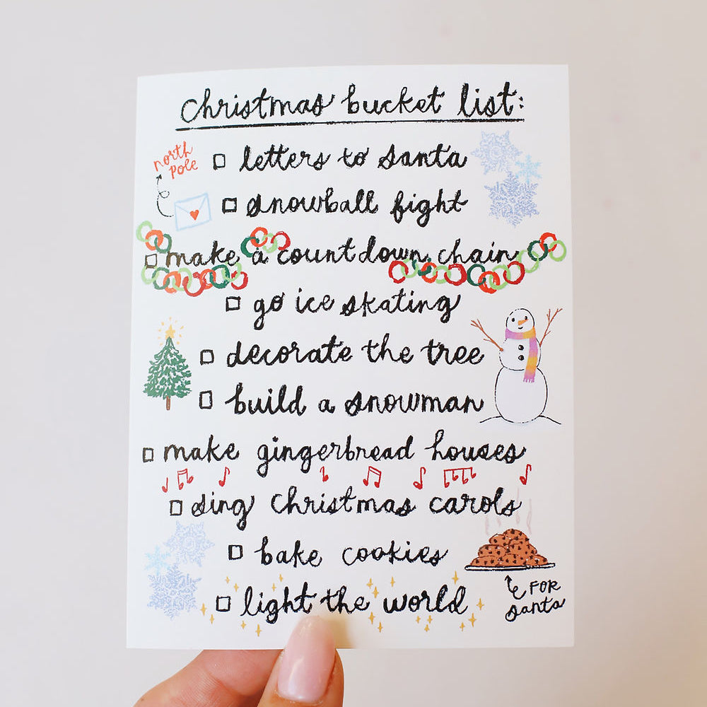 White card with a Christmas bucket list written in charming cursive with whimsical doodles of Christmas festivities nestled around the front of the card. Pictured in a woman's hand with a white back ground.
