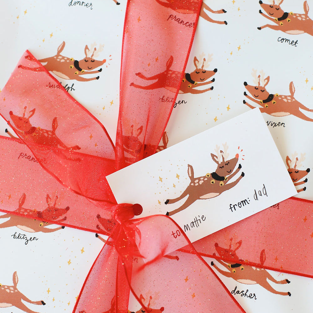 Reindeer tag pictured atop a red bow on a gift wrapped in matching reindeer wrapping paper.