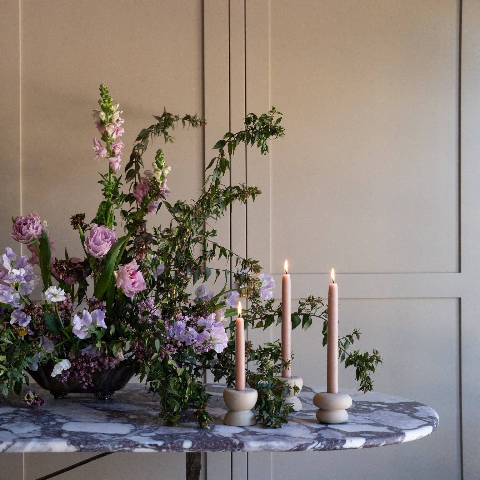 A styled floral arrangement with green foliage and pink and lavender flowers on a marble-topped table, alongside three lit taper candles in matte holders against a beige paneled wall.