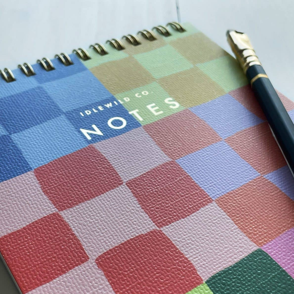 Colorful checkered notebook with 'Idlewild Co. Notes' text and a pen on a white background