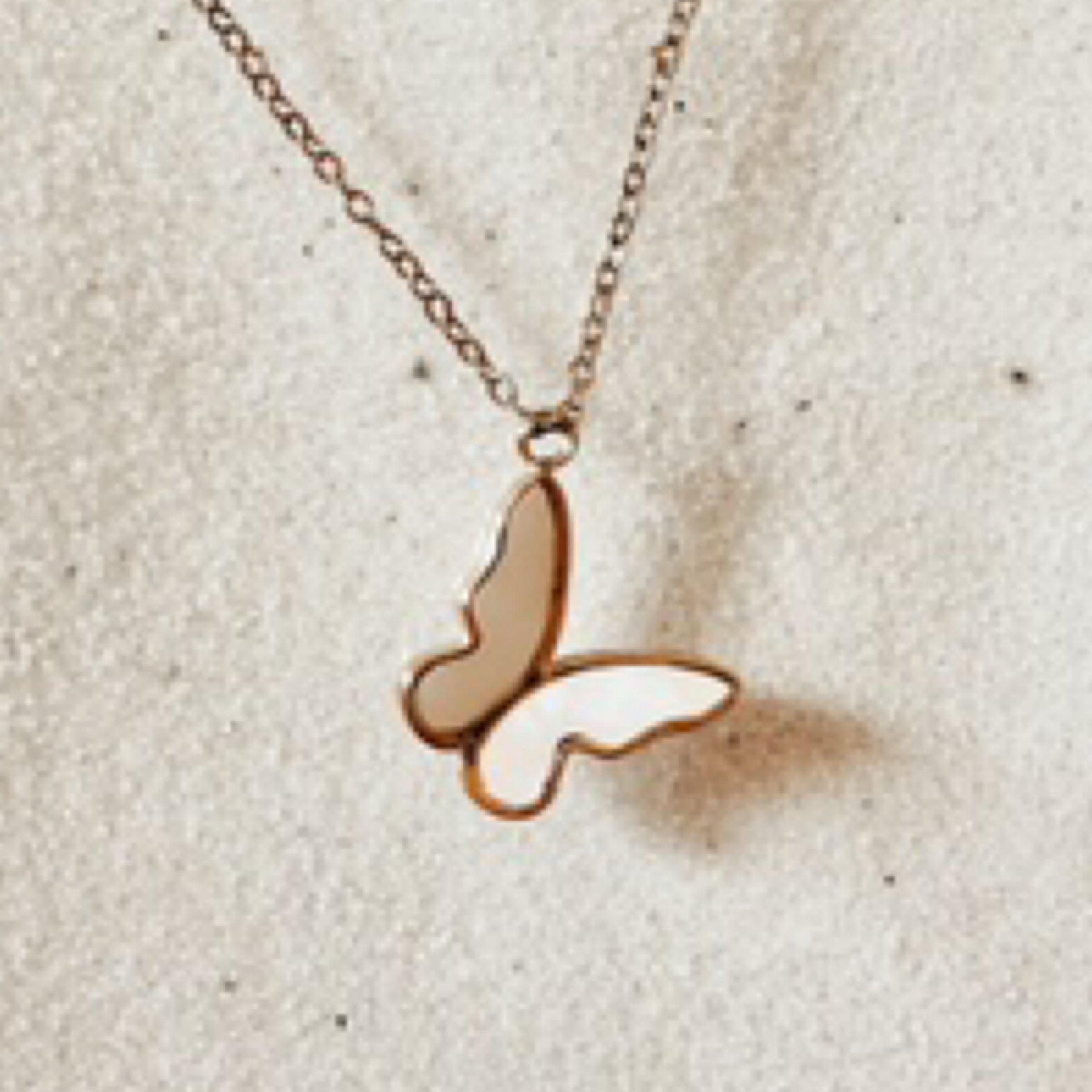 Delicate gold chain with a shimmery white butterfly outlined in gold. Pictured against white sand.