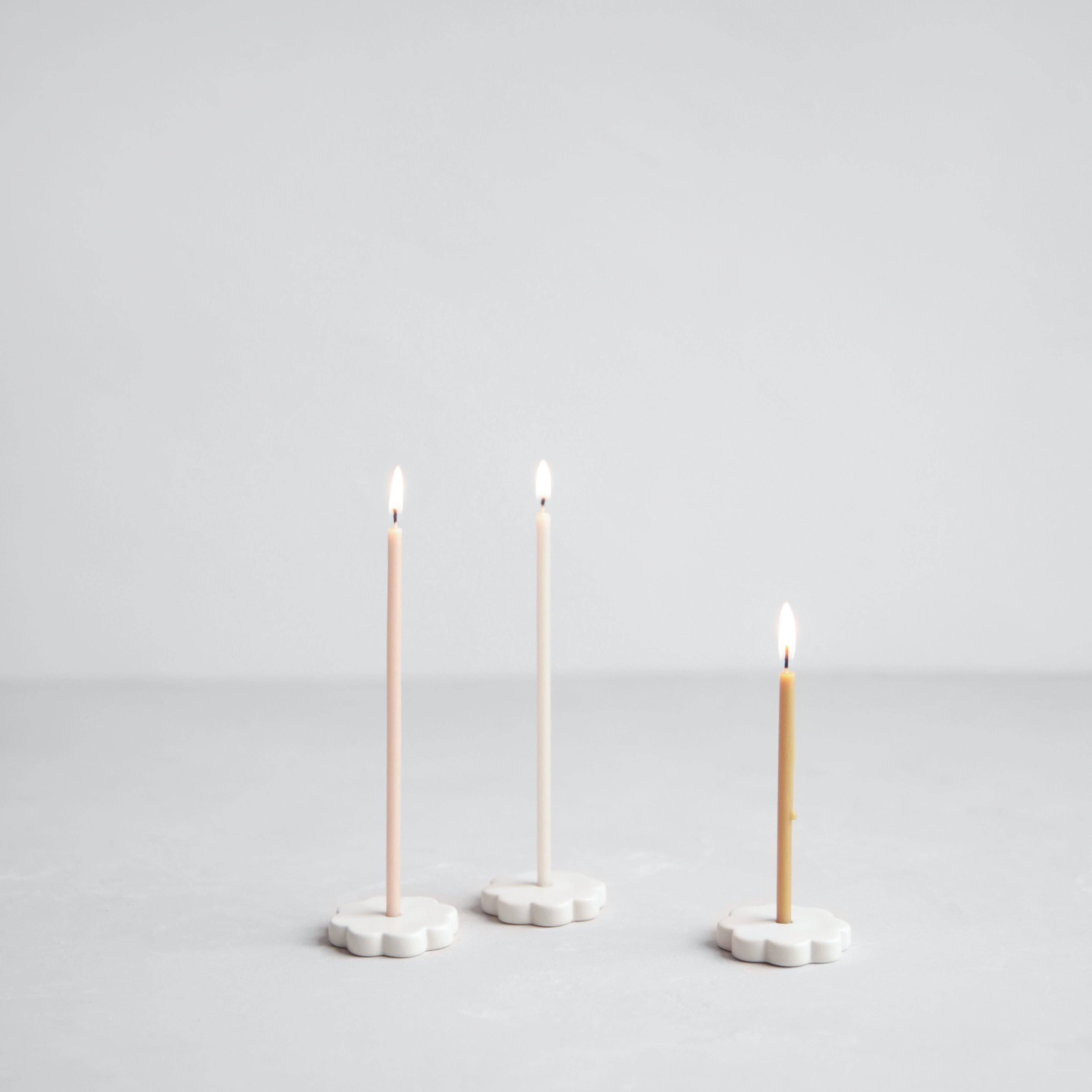 Three slim taper candles in soft blush, cream, and tan tones, each mounted on a small white flower-shaped base and lit, set against a seamless light gray studio background.