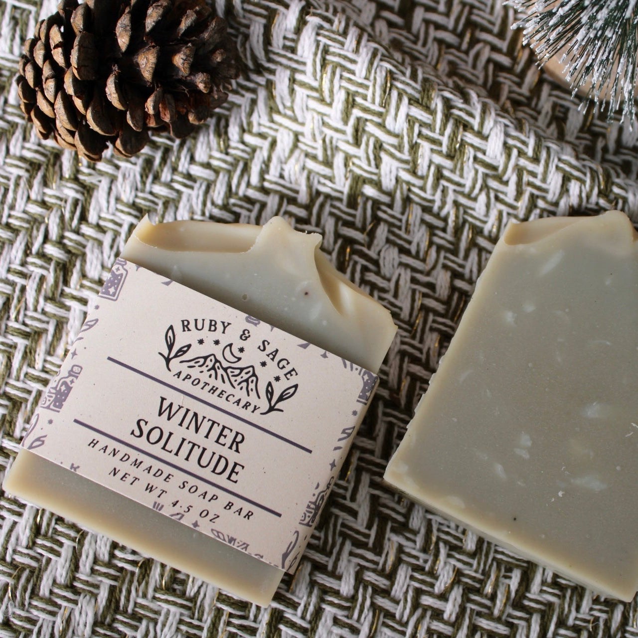 Light green bar soap with a cream label designed with sprigs and various fonts. Pictured alongside an unlabeled bar (for viewer to see full bar, with is flecked with white.) Pictured against a white and dark green weave with pinecones and frosted pines.