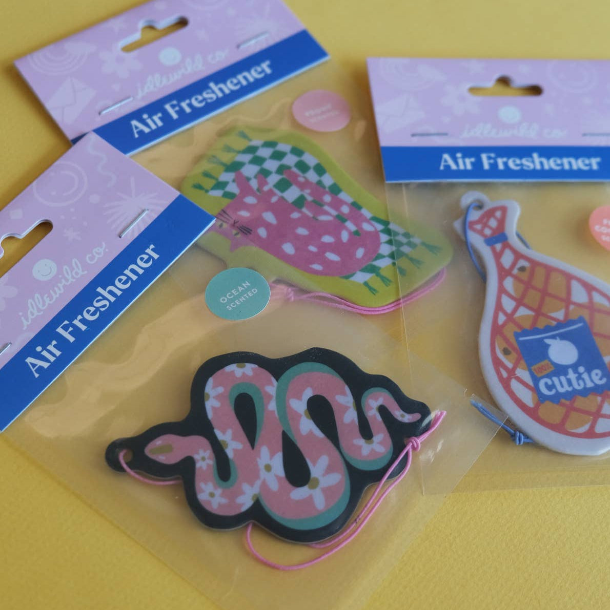 Colorful cat-shaped air freshener with a pink cat on a green and white checkered rug, attached to a pink string.
