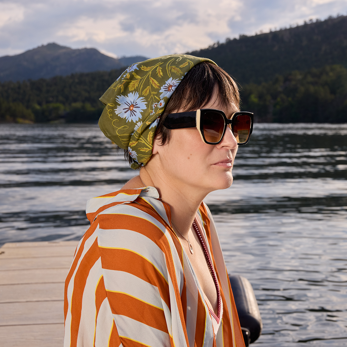 Woman wearing sunglasses and a patterned headscarf by a lake with mountains in the background