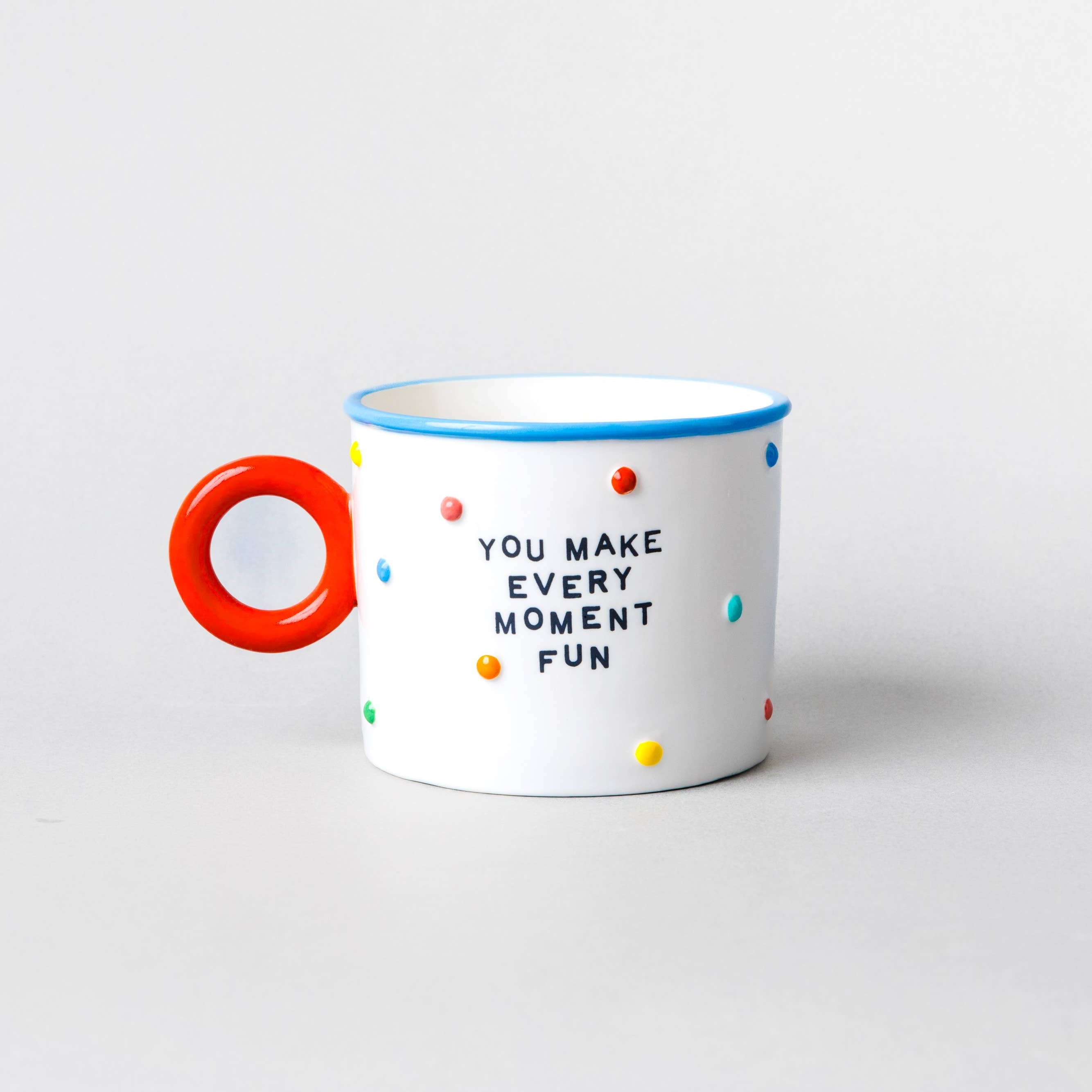 A white ceramic mug with a bright red handle, featuring the text 'YOU MAKE EVERY MOMENT FUN' in black lettering, decorated with small multicolored polka dots, and finished with a blue rim.