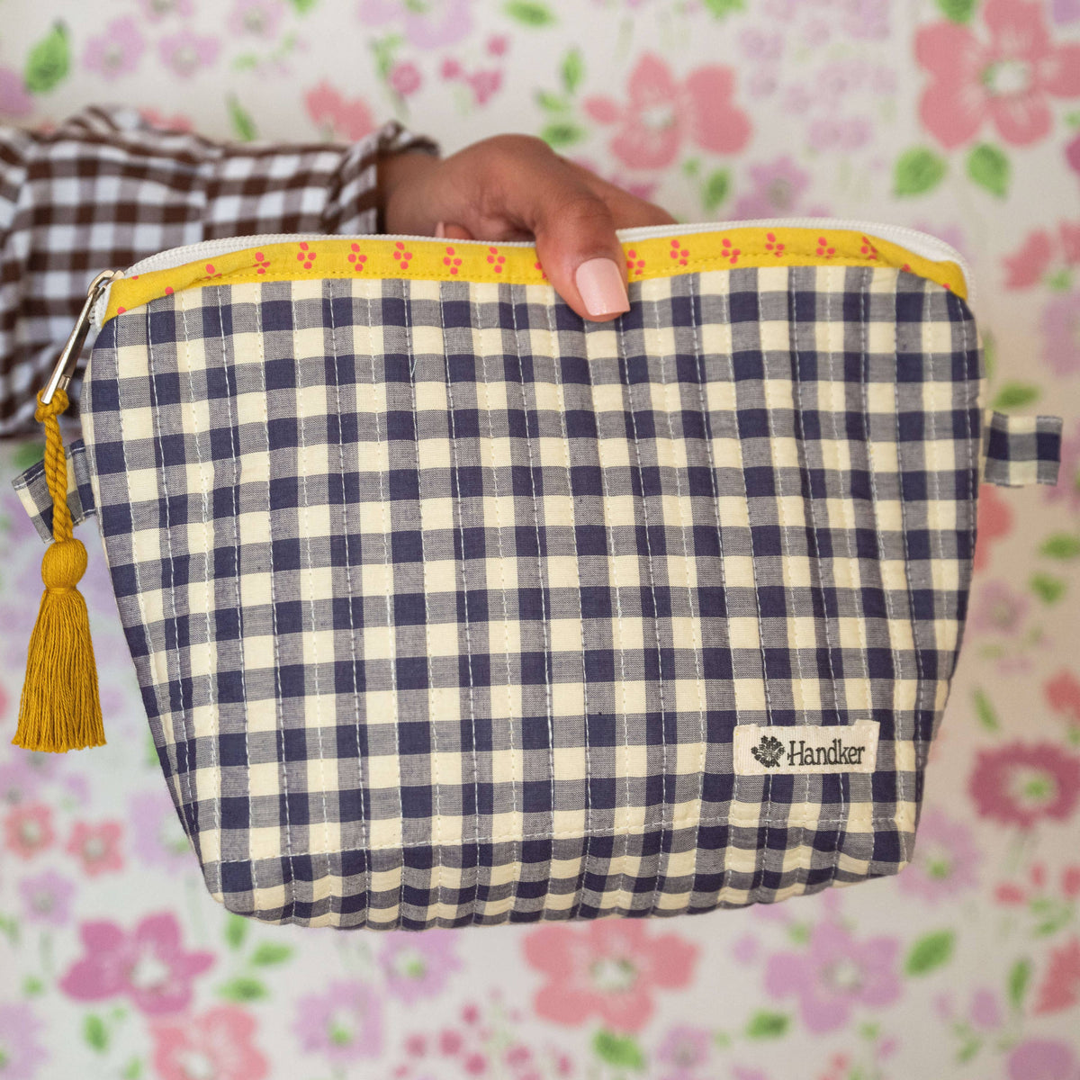 Hand holding a blue and white checkered pouch with a yellow zipper against a floral background
