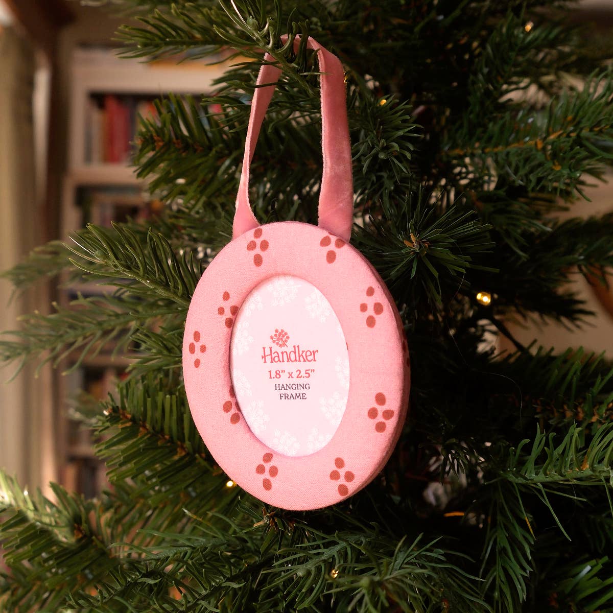 A pink fabric oval hanging frame ornament decorated with small red dots, hanging from a green Christmas tree branch. The center has a white insert with the brand name 'Handker' visible.