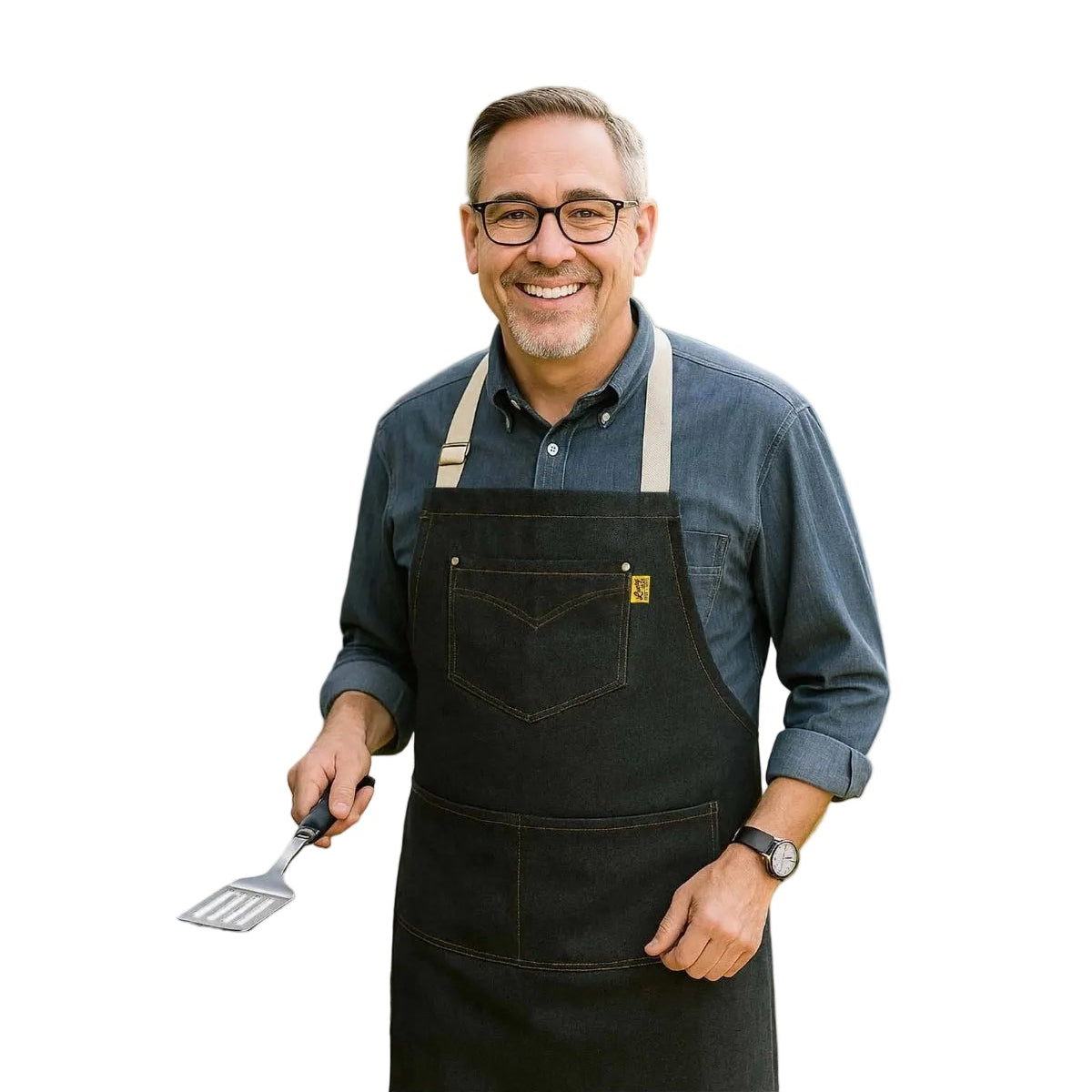 Man grilling outdoors wearing a black apron with a grill in the foreground and people in the background.