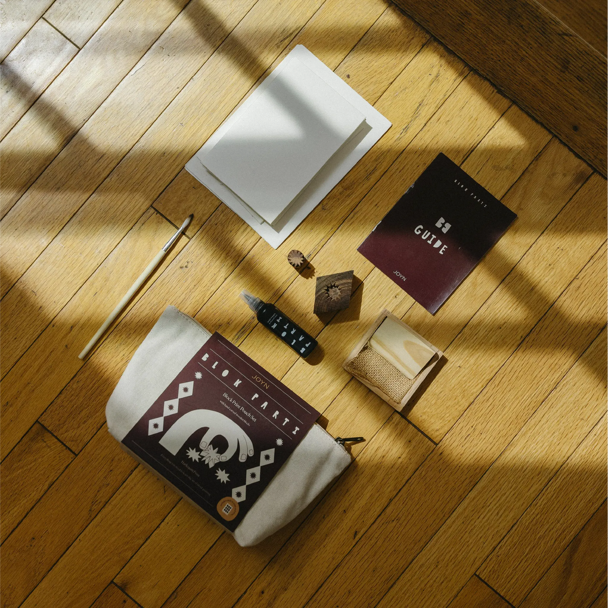 A styled flat-lay of a desk accessories set on a wooden floor with dramatic window shadows. The set includes a cream canvas pencil pouch with a dark maroon printed cover, a white notebook, a dark maroon guidebook, a black pen, a small wooden cube, a wooden pencil, and a small beige pouch.