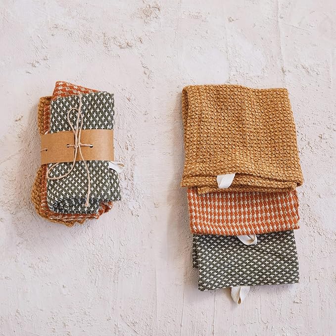 Folded dish towels in brown, green, and orange on a textured white surface