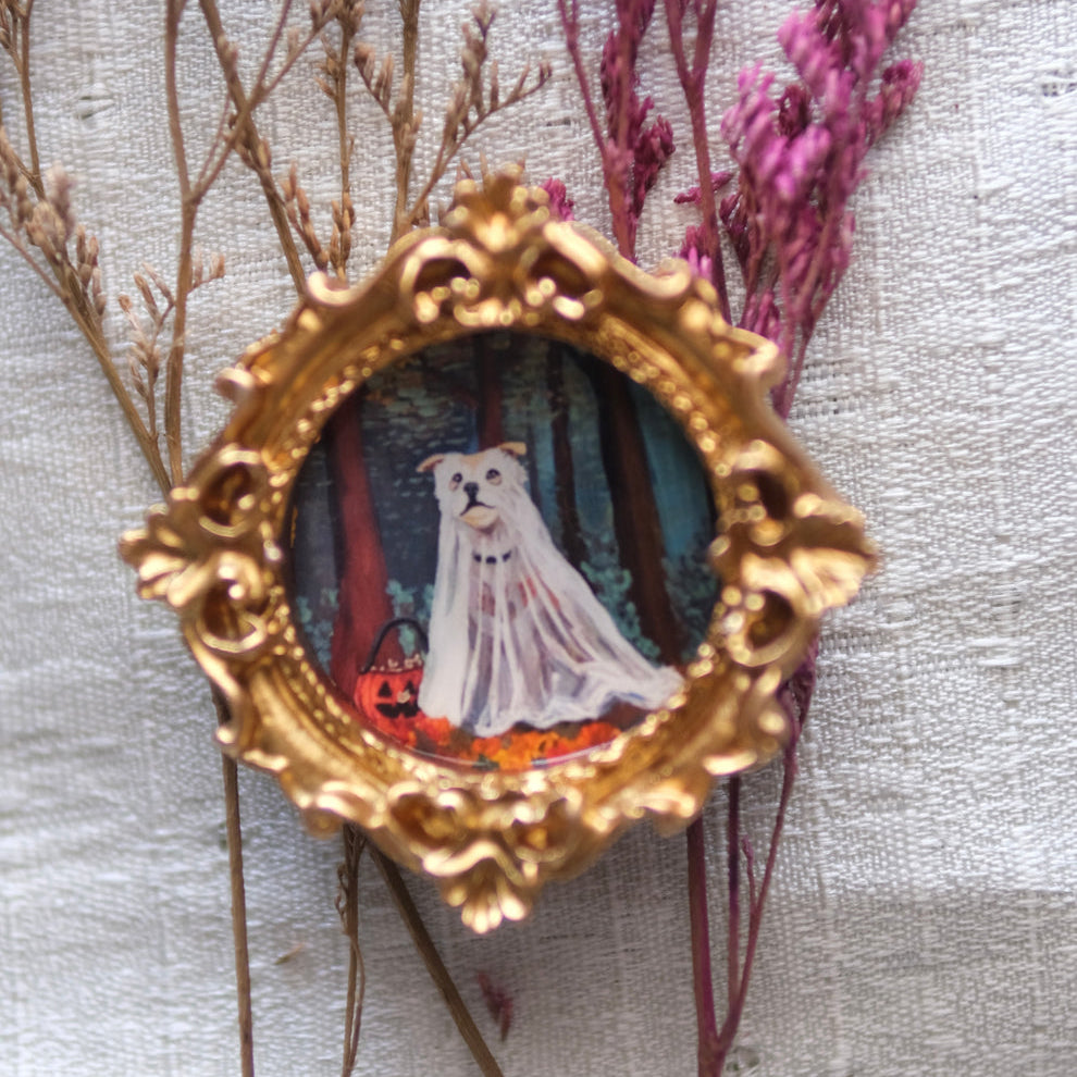 A small ornate gold frame encloses an illustration of a white dog dressed as a ghost in a sheet, standing in a dark forest with a jack-o'-lantern basket nearby. The frame is nestled among dried beige and purple branches on a soft white fabric background.