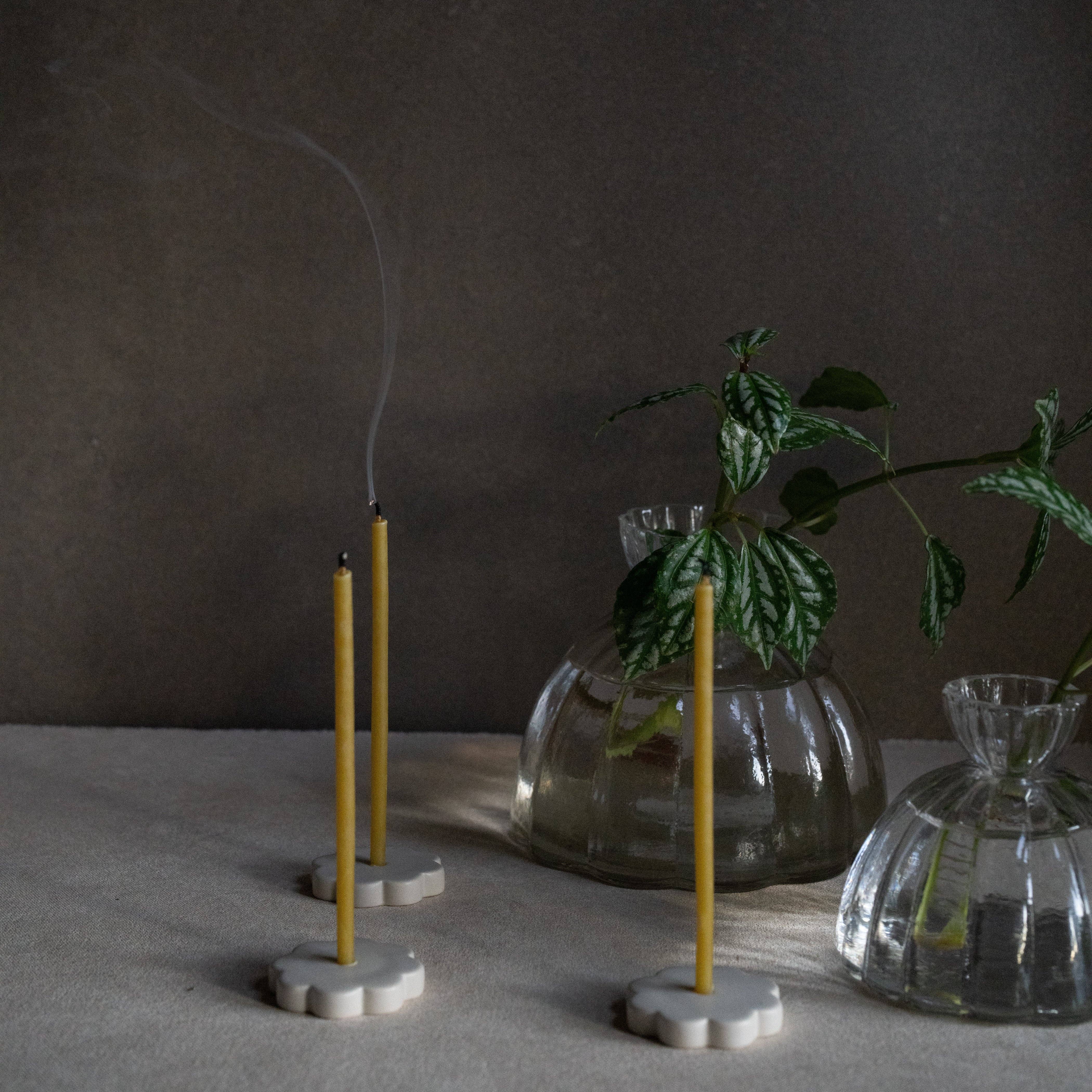 Three yellow taper candles in white flower-shaped holders on a neutral surface, with one candle lit and smoke curling upward. A glass vase with green leaves and a small clear glass container sit beside them against a dark brown background.