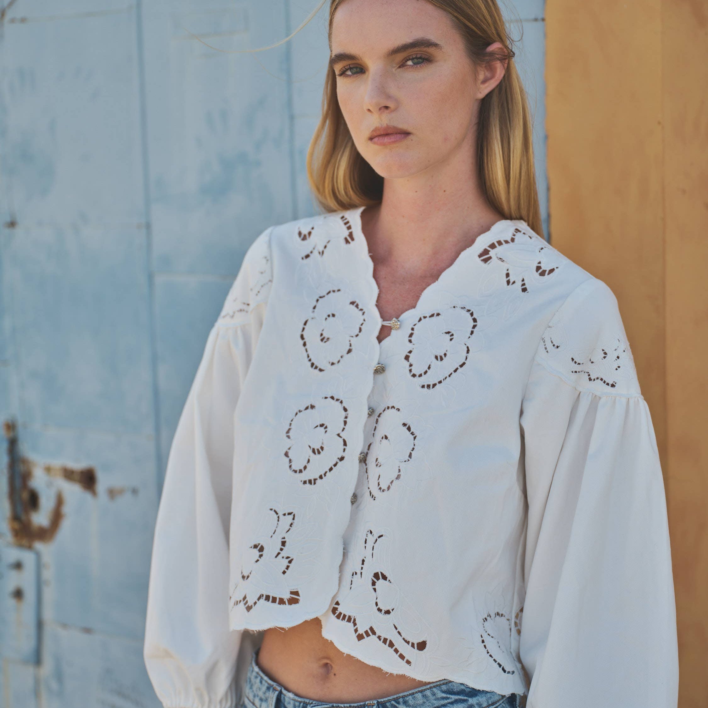 Woman wearing a white embroidered blouse and blue jeans against a rustic wall.