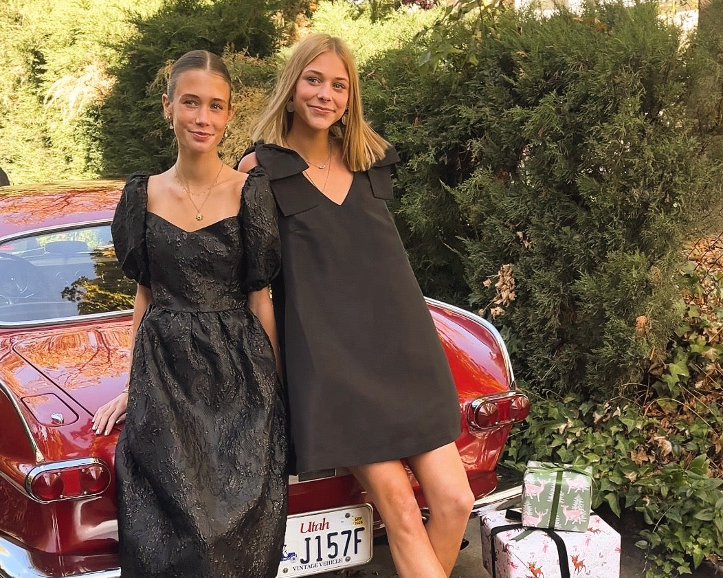 Two women in black dresses standing next to a vintage car with gift boxes nearby.