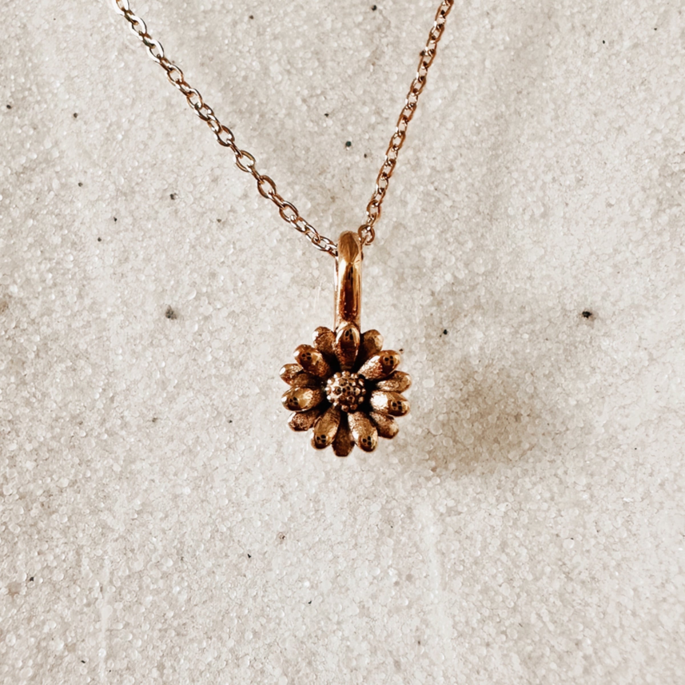Delicate gold chain with a detailed gold daisy pendant. Pictured against white sand.